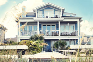 Seaside Park vacation rental blue with white trim 3-story; back of home is shown surrounded by beach grass in the landscape